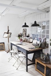 a whiet Scandinavian home artist studio with a shared desk, a couple of chairs, black pendant lamps, chests and crates and an easel