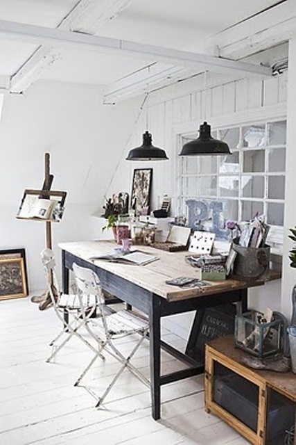 a whiet Scandinavian home artist studio with a shared desk, a couple of chairs, black pendant lamps, chests and crates and an easel