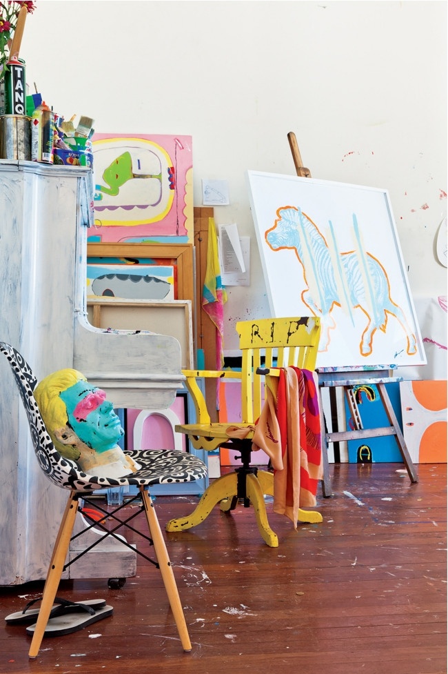 A bright home studio with a whitewashed piano, some shelves, an easel, a chair, colorful artwork and paints