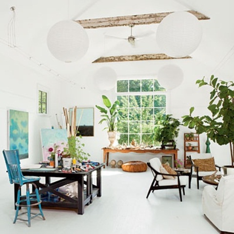 a white light-filled home artist studio with wooden beams, tables and chairs, a sitting zone, potted plants and artwork