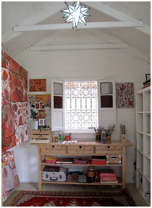 a small artist home studio with a window with shutters, colorful artwork, a wooden desk with storage shelves and a bold rug