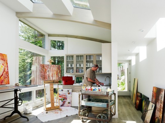 a beautiful light-filled artist home studio with skylights and windows, glass front cabinets and usual ones with paints, brushes and other supplies, an easel and a cart