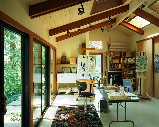 A mid century modern home studio with glazed walls and skylights, a large table and a black chair, bookshelves and some easels with artwork