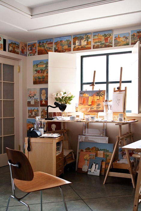a small and catchy home artist studio with a trestle desk, a chair, a ledge with artwork, a storage cabinet and a table for storing supplies