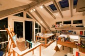 an attic light-filled home artist studio with a glazed ceiling, walls and windows, with several tables or desks, an easel, baskets and lots of paints