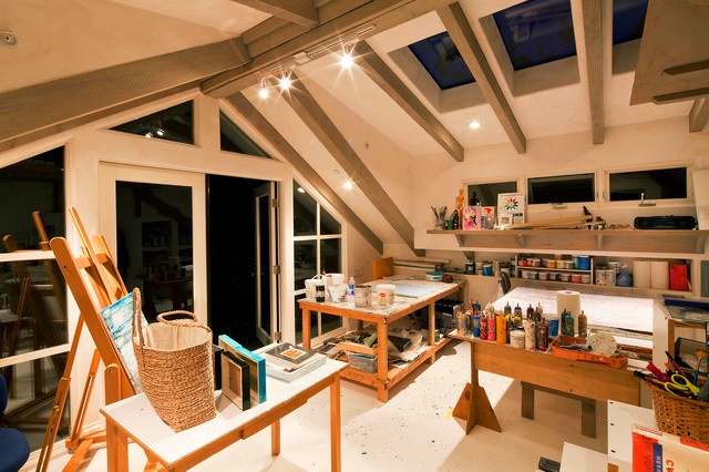 An attic light filled home artist studio with a glazed ceiling, walls and windows, with several tables or desks, an easel, baskets and lots of paints