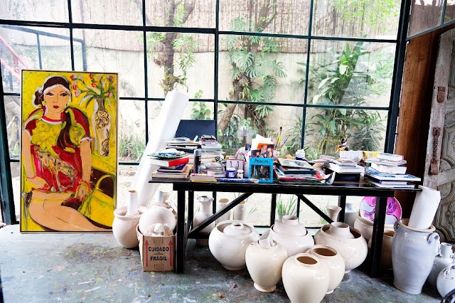 A light filled home artist studio with a glazed wall, a large desk, pottery with paper and a large artwork is a cool space