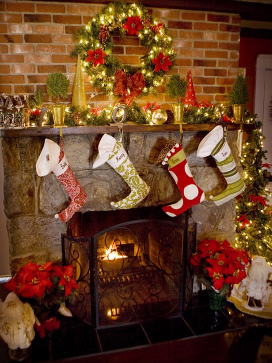 a traditional Christmas mantel with en evergreen garland with lights, potted mini trees and a bold Christmas wreath with red blooms and stockings hanging over the fireplace