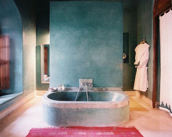 a blue plaster bathroom with a concrete bathtub, wooden shutters and a pink rug
