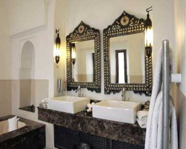 a monochromatic Moroccan bathroom with a stone vanity, ornated mirrors and wall lamps