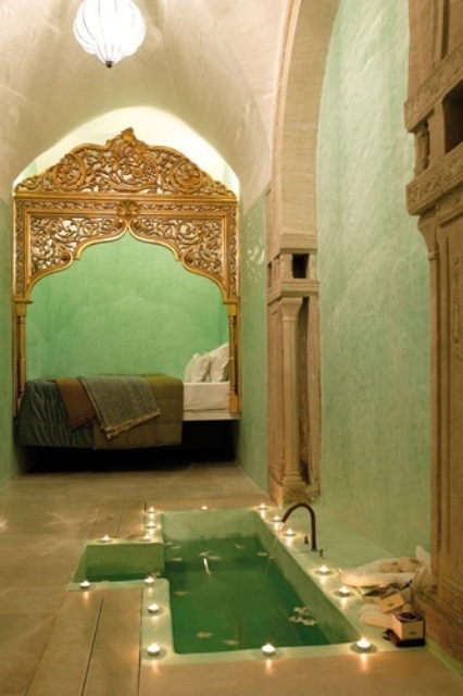 a grene bathroom located in a bedroom, with a sunken bathtub with candles and a faucet plus pillars built in the walls