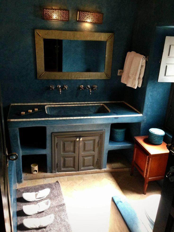 a navy Moroccan bathroom with a built-in vanity, a gilded frame mirror, wall lamps and a wooden sideboard