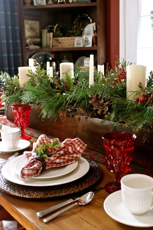 a lush rustic Christmas tablescape with woven placemats, red glasses, a box with evergreens, pinecones and candles