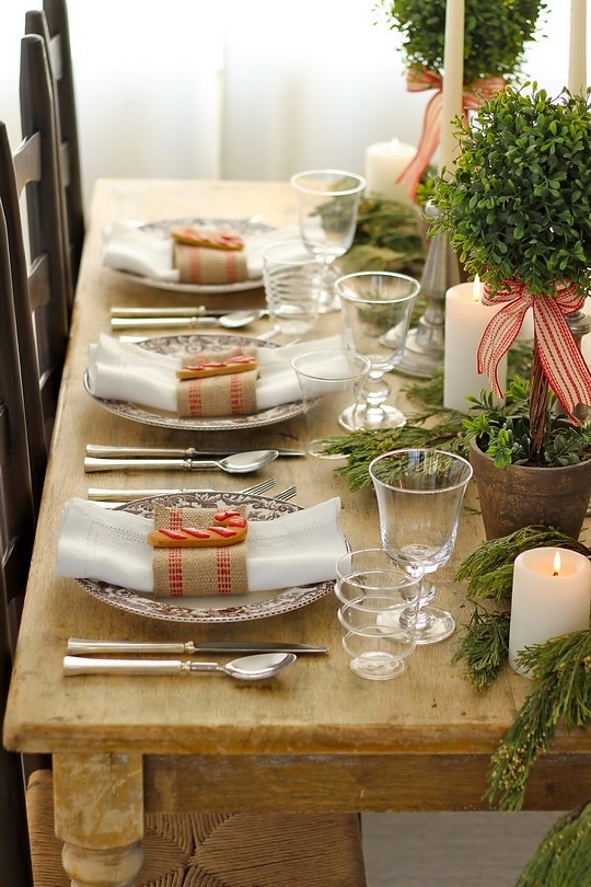 a rustic Christmas tablescape with an uncovered table, evergreens in pots, candles, refined chargers and striped napkins