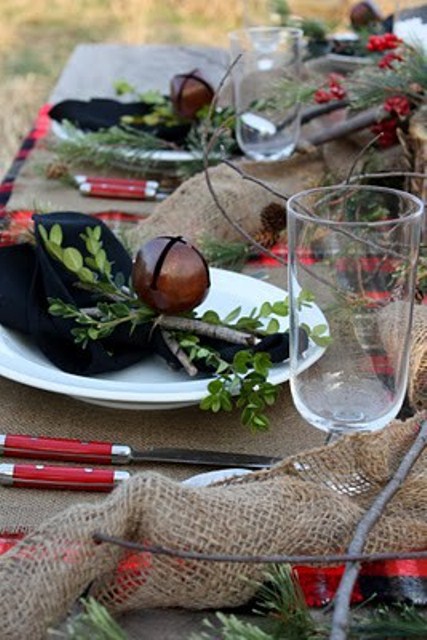 a rustic Christmas table setting with a burlap tablecloth and napkins, berries, evergreens, bells and sticks for a natural feel