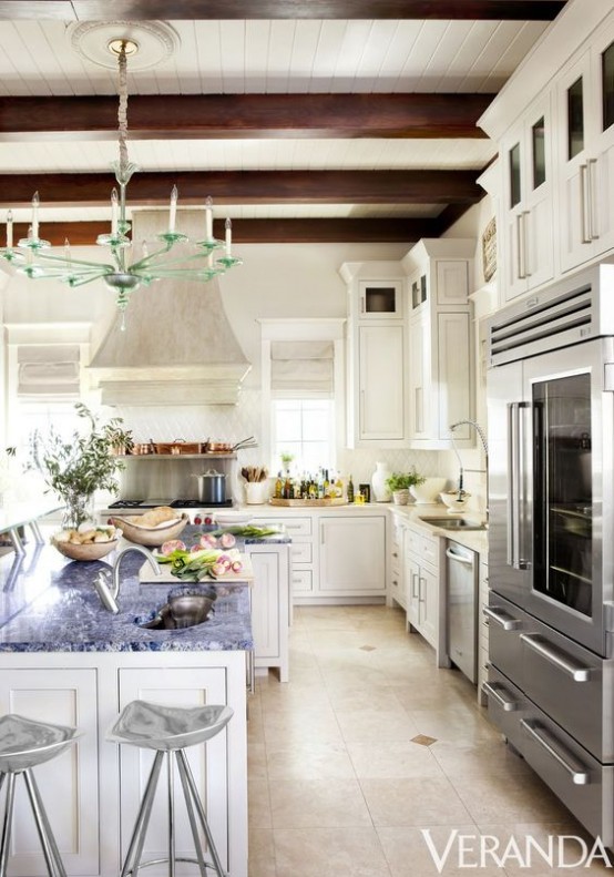 an elegant white vintage kitchen with chic cabinetry, a large kitchen island, chic stools, dark wooden beams for a refined feel and a creative glass chandelier