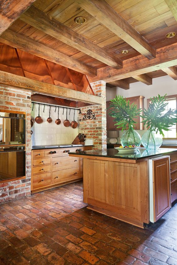 A cozy warm tone kitchen with wooden cabinetry, brock walls, black stone countertops, wooden beams on the ceiling to add interest and an architectural element to the space