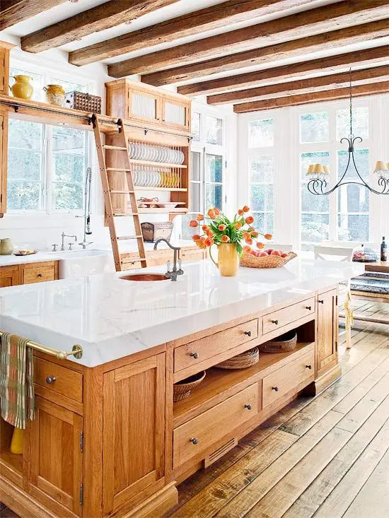 A warm colored farmhouse kitchen with rich colored cabinetry, white marble countertops, wooden beams on the ceiling and a vintage chandelier