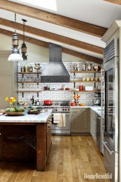 a cozy farmhouse kitchen with neutral cabinetry and stainless steel appliances, a dark heavy kitchen island, wooden beams that highlight the attic ceiling and vintage lamps hanging on them