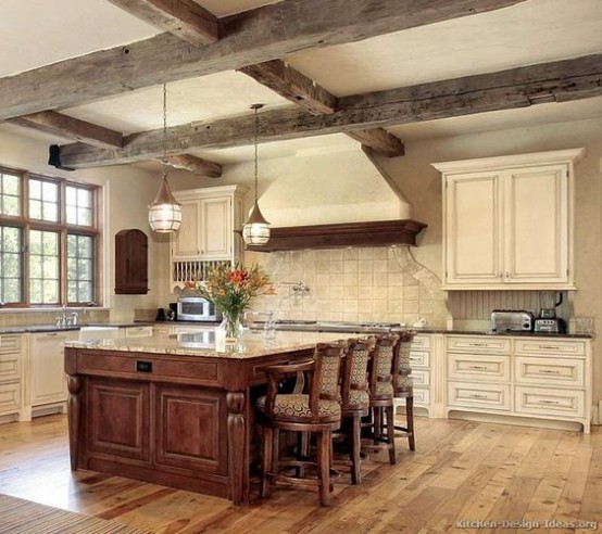 a warm-colored farmhouse kitchen with vintage cabinetry, a large hood, a dark carved kitchen island and exposed wooden beams with pendant lamps is very refined