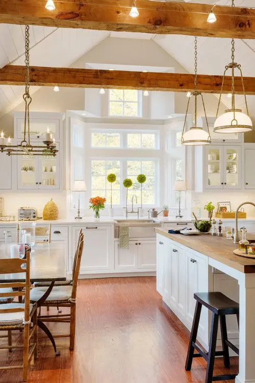 a white modern farmhouse kitchen with chic cabinetry, wooden beams with lights and elegant chandeliers and lamps