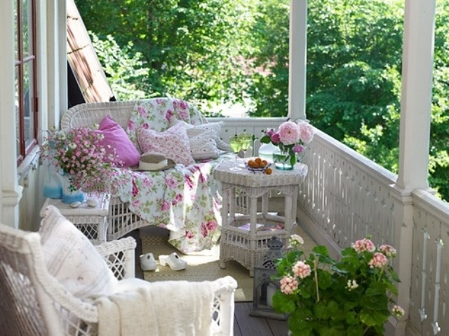 A vintage inspired summer porch with white wicker furniture, bright and floral print textiles, potted blooms and candle lanterns
