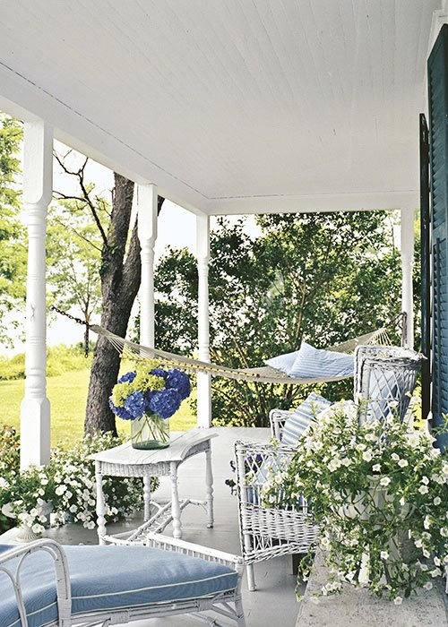 a pastel and neutral porch with wicker furniture, a hammock, potted blooms and greenery