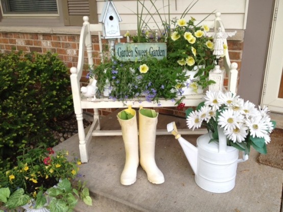 a small porch decorated with a vintage bench with potted blooms, a sign, a watering can with blooms and rubber boots