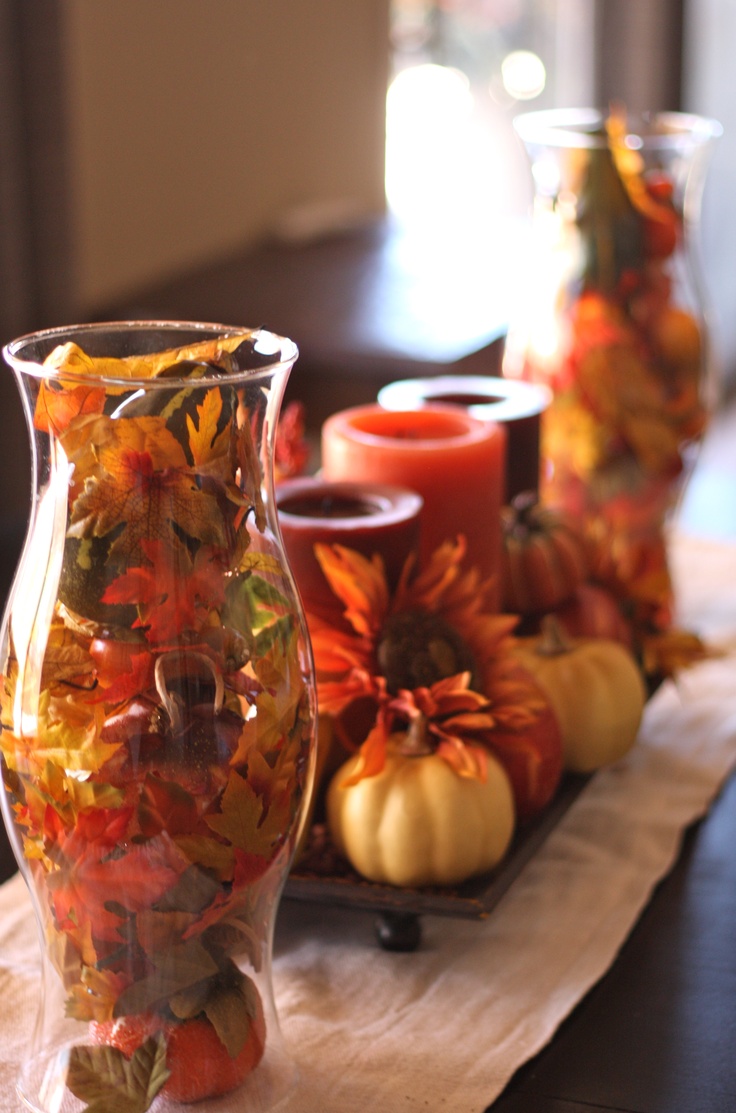 A clear vase with faux fall leaves and berries plus a stand with faux blooms, pumpkins and fall colored candles