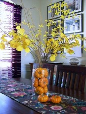 a bright and simple fall centerpiece of a clear vase filled with mini pumpkins and branches with yellow leaves
