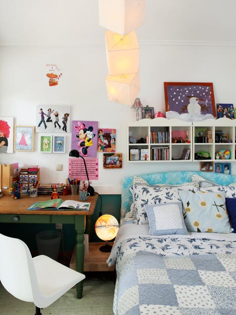 A small and pretty girl's room with a bed with printed bedding, box shaped shelves, a bright gallery wall, a desk and a creamy chair, paper lights