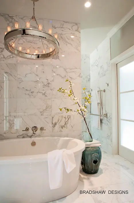 a luxurious modern bathroom clad with white marble, with a round tub, a statement chandelier and a frosted glass window