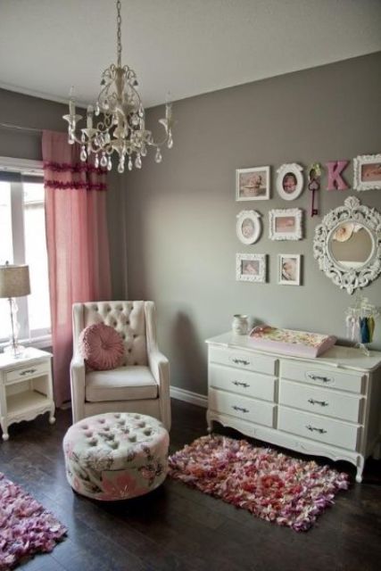 a vintage nursery with grey walls, a creamy chair and a footrest, a white dresser, a crystal chandelier and pink textiles - curtains and rugs