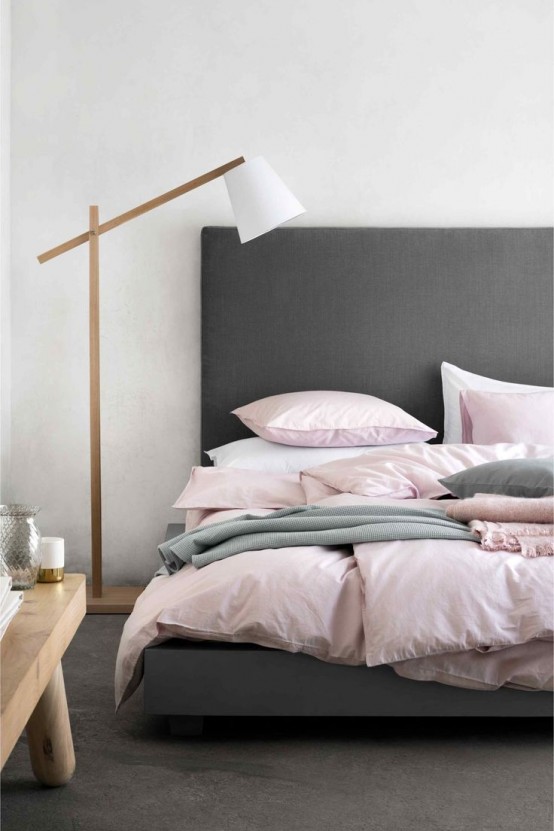 a laconic bedroom with a graphite grey bed with pink and grey bedding, a floor lamp and a wooden bench