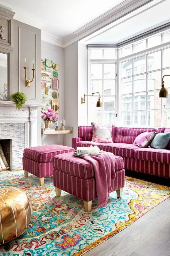 an adorable vintage living room with a large bow window, grey walls and paneling, a hot pink sofa and poufs, a bright blue and gold rug