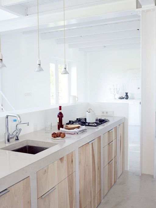 A minimalist light filled kitchen with wooden cabinets and white concrete countertops plus pendant bulbs