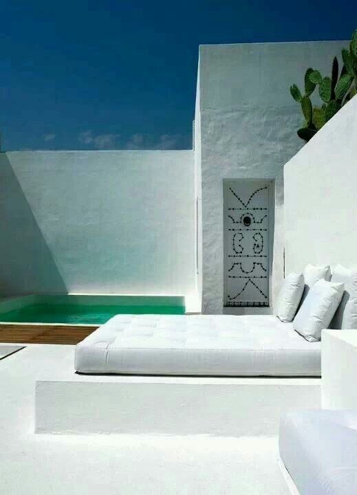 A minimalist white terrace with a small pool, a built in bed and seats, a decorated niche and a mattress with pillows is a very laconic space