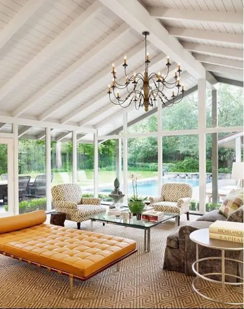 a modern sunroom with glazed walls, a taupe sofa, printed chairs, a coffee table, an orange leather daybed and a chic vintage chandelier