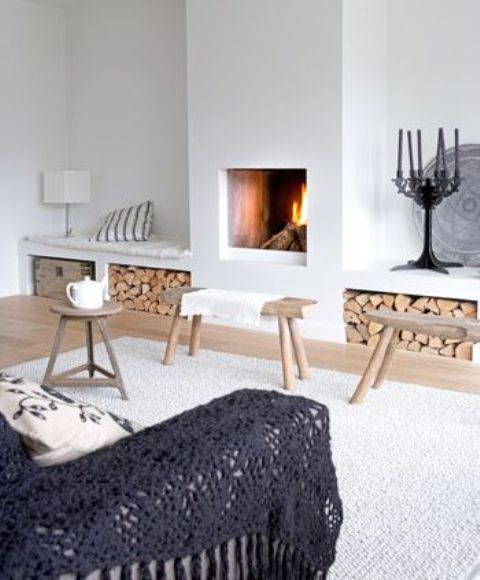 A stylish and welcoming Scandinavian living room with a built in fireplace and firewood storage niches that double as benches, a sofa with a dark cover and some small stools and benches