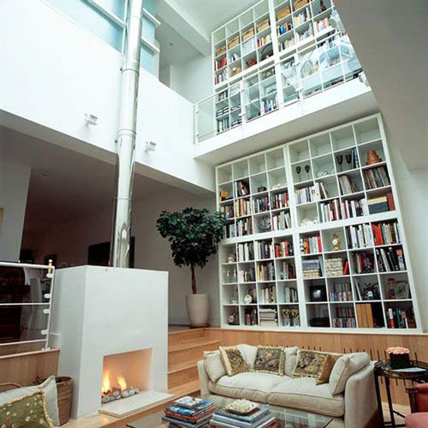 A double height white library with lots of bookshelves, a sleek white fireplace and some neutral sitting furnitureplus skylights over it