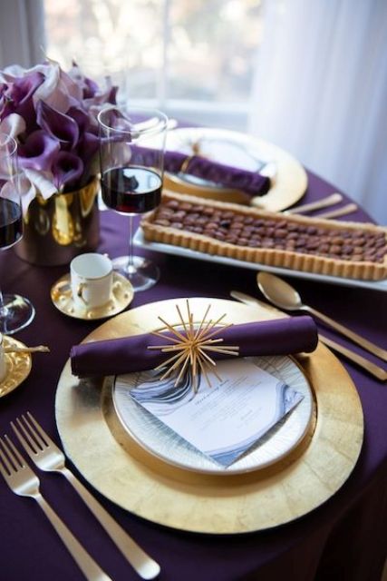 a bold modern Thanksgiving tablescape with a purple tablecloth and napkins, gold chargers, cutlery and saucepans and deep purple blooms