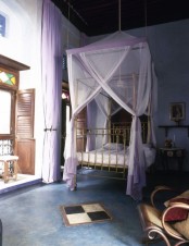 a Moroccan bedroom with a gold bed and lavender canopy, shutters and a mosaic balcony entrance, carved wooden furniture