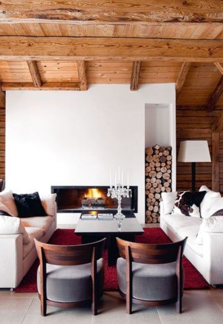 A bold chalet living room with a wooden ceiling with beams, a built in fireplace, white sofas and low grey chairs is a welcoming and cool space