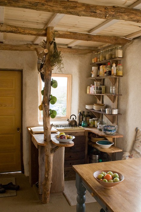 a small wabi-sabi kitchen with open shelves, rough wooden beams on the ceiling and a pillar holding cups, an additional table that can be used as a kitchen island