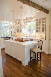 a white farmhouse kitchen with shaker and glass cabinets, a wooden beam, crystal chandeliers and a vintage chair
