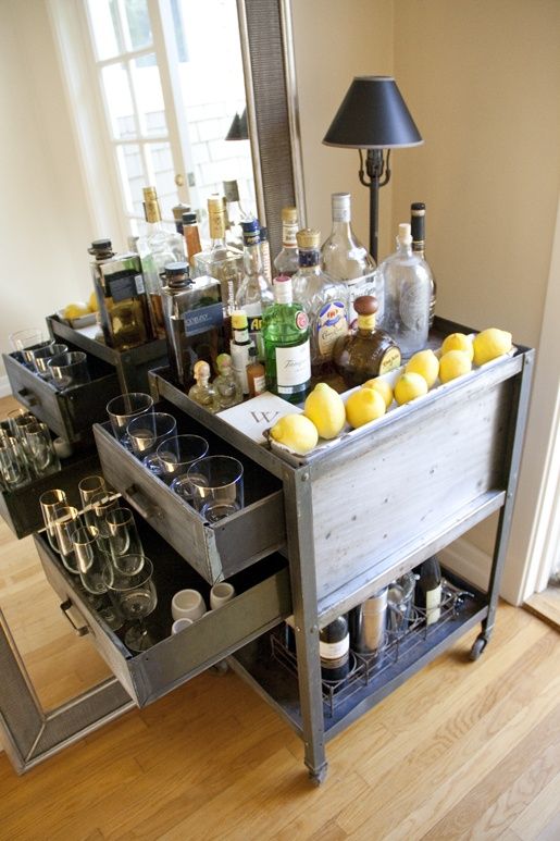 a small industrial home bar of aged metal, with drawers and casters, with a table lamp and lots of bottles, glasses and lemons is a stylish piece that you can DIY