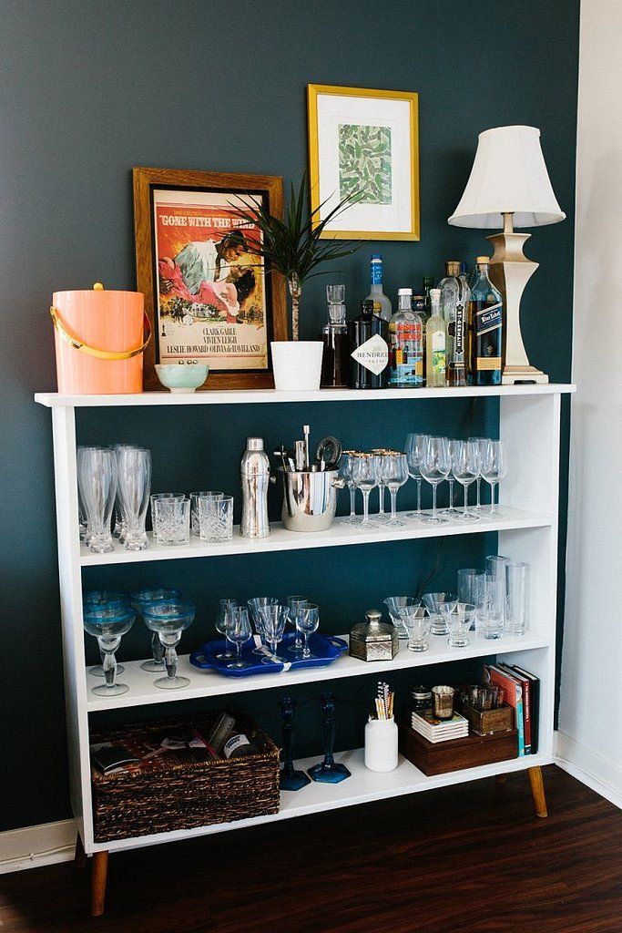 A mid century modern shelving unit in white turned into a home bar, with baskets, lots of glasses and wine bottles, decor and a table lamp