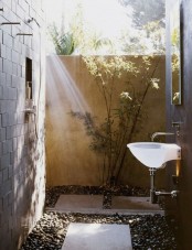 a simple outdoor shower with pebbles and tiles on the ground, greenery and a sink