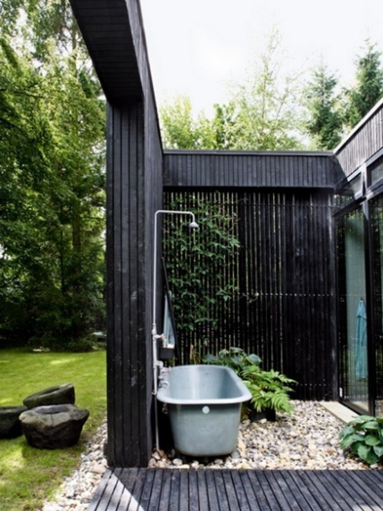 a moody outdoor bathroom with black wooden plank walls, a blue bathtub and pebbles on the wall