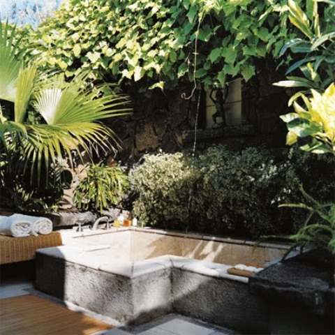 a refined outdoor bathroom with a concrete bathtub, a bench with towels and greenery